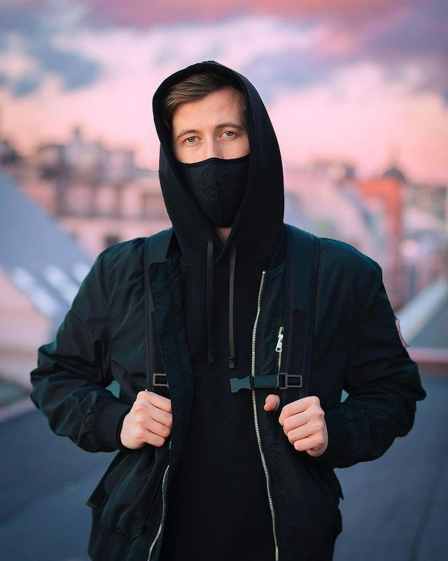Young man in black hooded streetwear and fabric face mask standing on a city rooftop at pastel su...