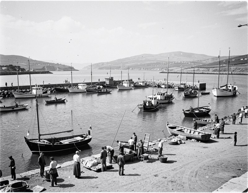 Vintage black and white view of fishing boats anchored in a coastal harbor with people gathered on the shore