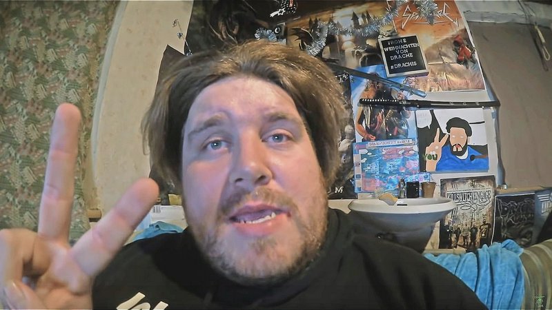 Bearded man holds up a peace sign to webcam, with a wall of band posters and fan art behind him