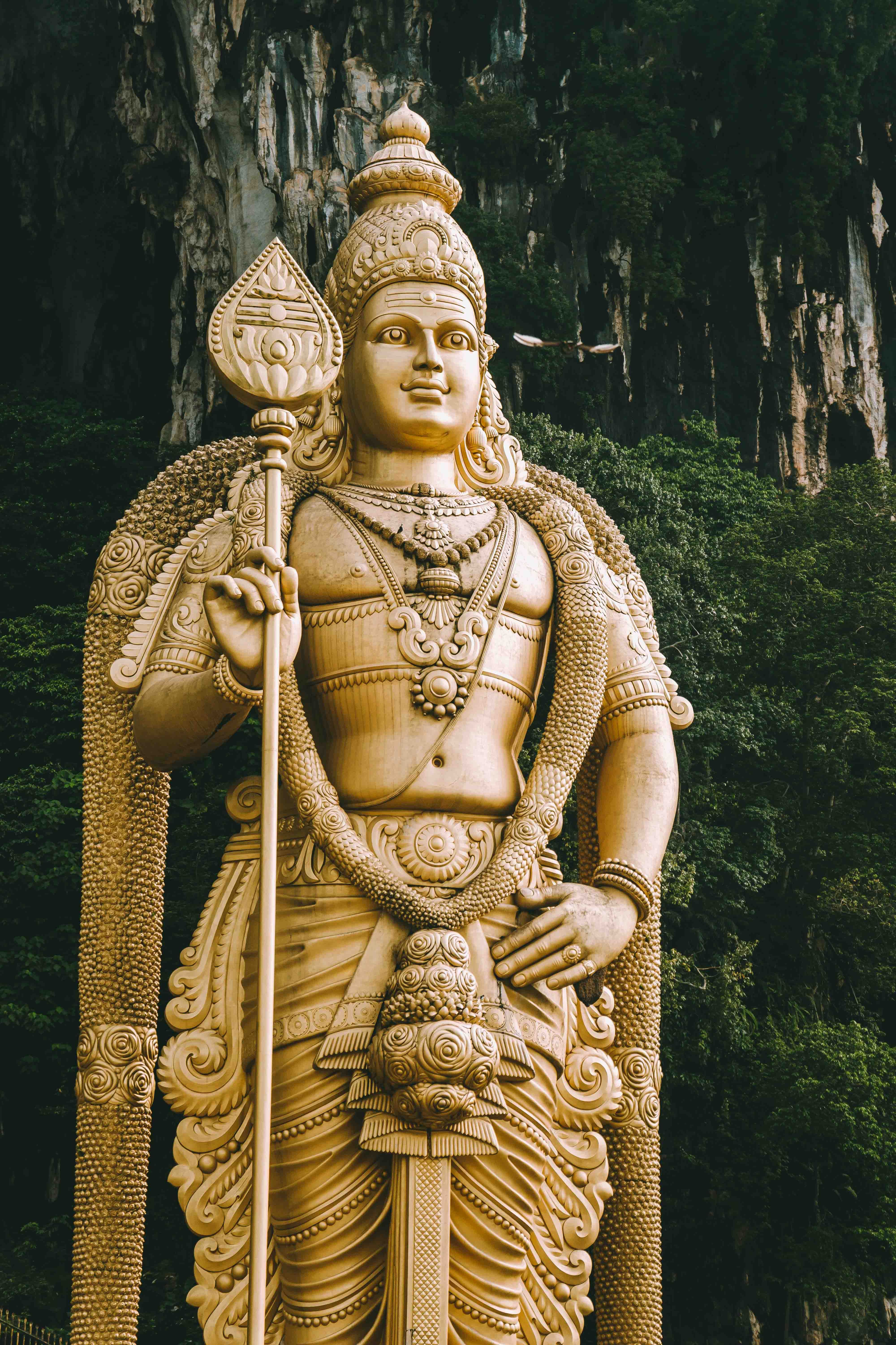 Tall gilded Lord Murugan statue stands before forested limestone cliffs at Batu Caves