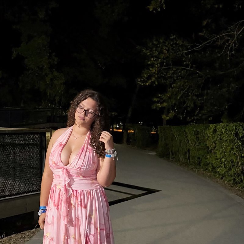 Young woman in a pink halter maxi dress poses on a paved park walkway at night