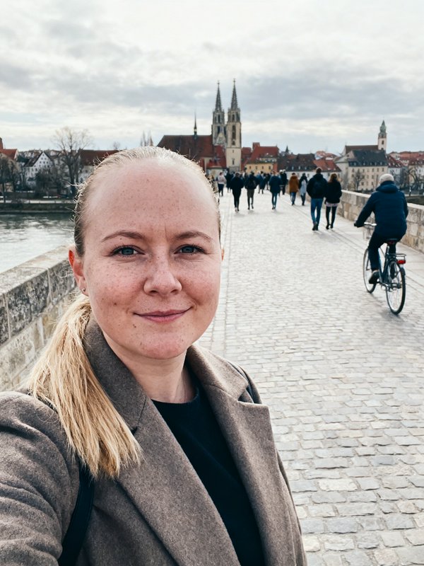 Woman taking a selfie on a cobblestone bridge with a European city and cathedral towers in the background.