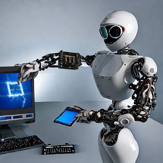 Sleek white humanoid robot holding a tablet while interacting with a desktop computer on a grey w...