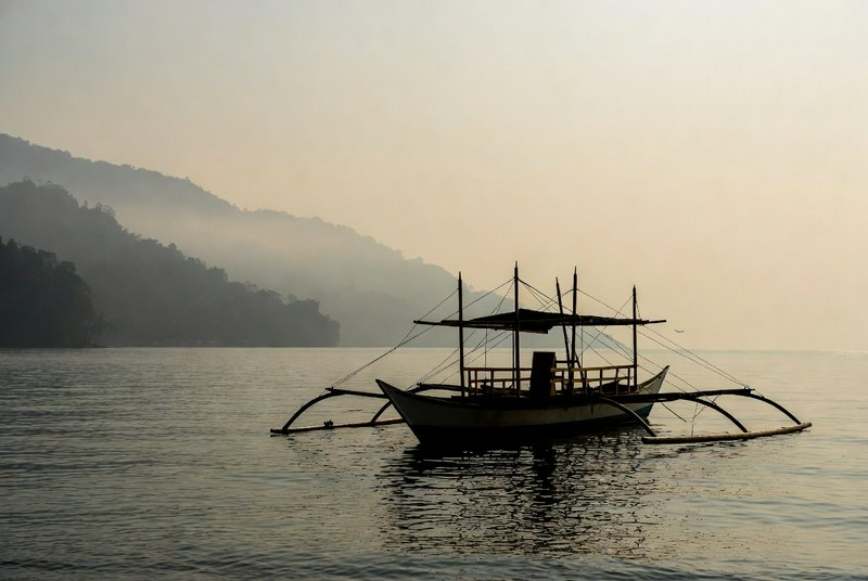 Silhouetted traditional boat with outriggers on calm water, hazy hills in the background.