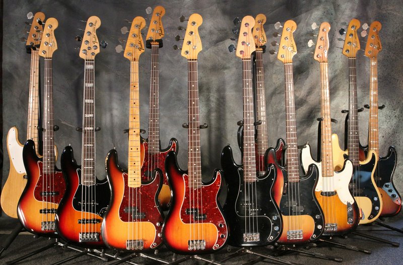 Row of assorted Fender Precision bass guitars displayed on stands against a grey studio backdrop