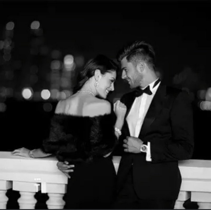 Elegant formally dressed couple smiling at each other on a night balcony with blurred city lights...