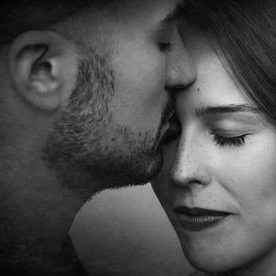 Intimate black and white close up of a bearded man gently kissing a woman's forehead as she rests...