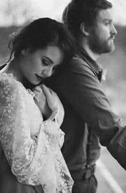 Soft monochrome portrait of a bride resting her head on her bearded partner's shoulder outdoors