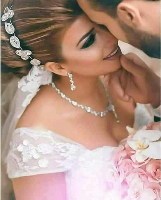 Groom tenderly kisses a bride's forehead as she wears sparkling bridal jewelry and holds a weddin...