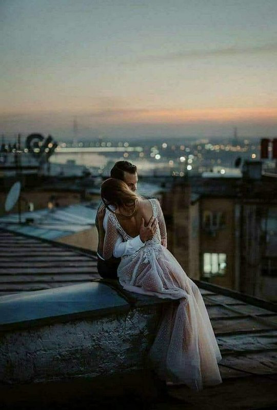 Bride and groom embrace while sitting on a city rooftop overlooking a glowing dusk cityscape