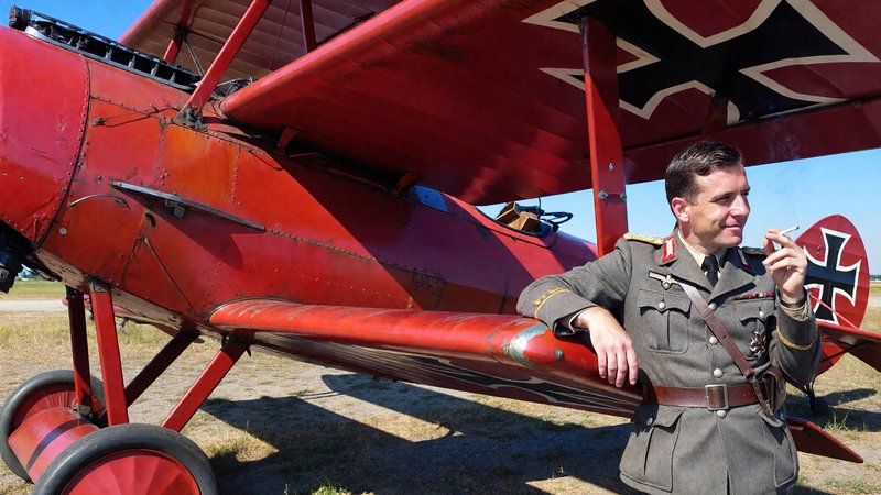 Reenactor in WWI German pilot uniform leans on the iconic red Red Baron Fokker triplane on a sunn...