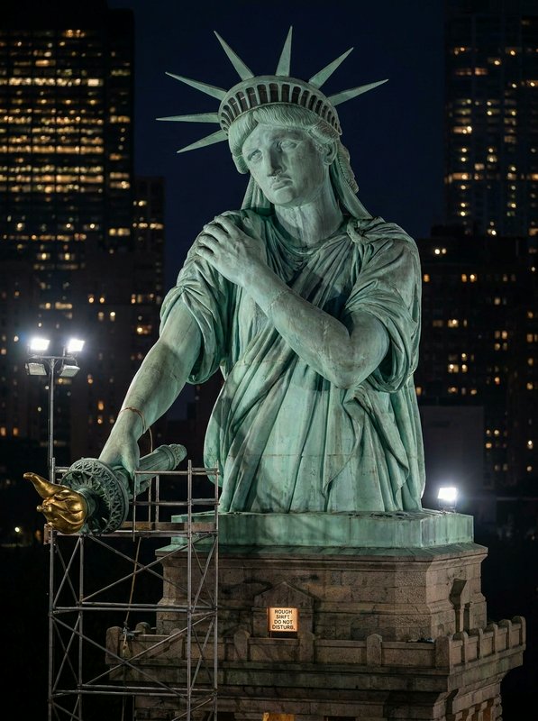 Nighttime view of the Statue of Liberty with scaffolding on the pedestal and city lights in the background.