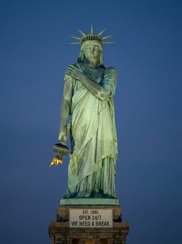 Statue of Liberty replica lit at dusk with a humorous plaque reading "WE NEED A BREAK" against a deep blue sky.