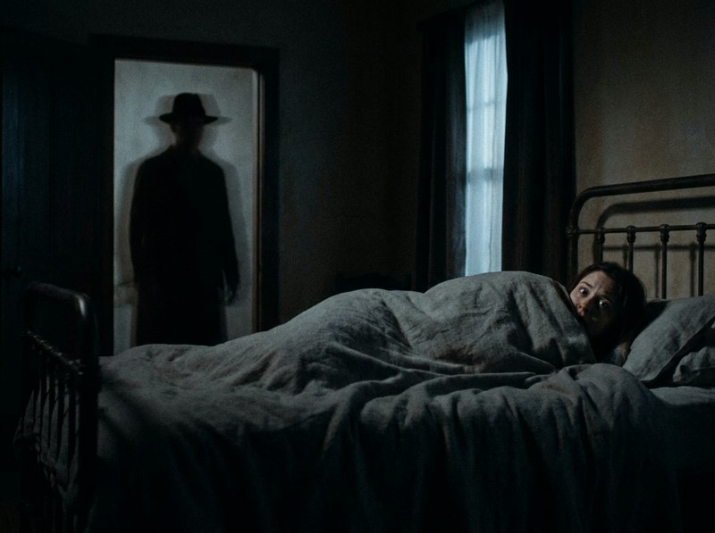 Woman in bed awake as a shadowy figure wearing a hat appears in the doorway.