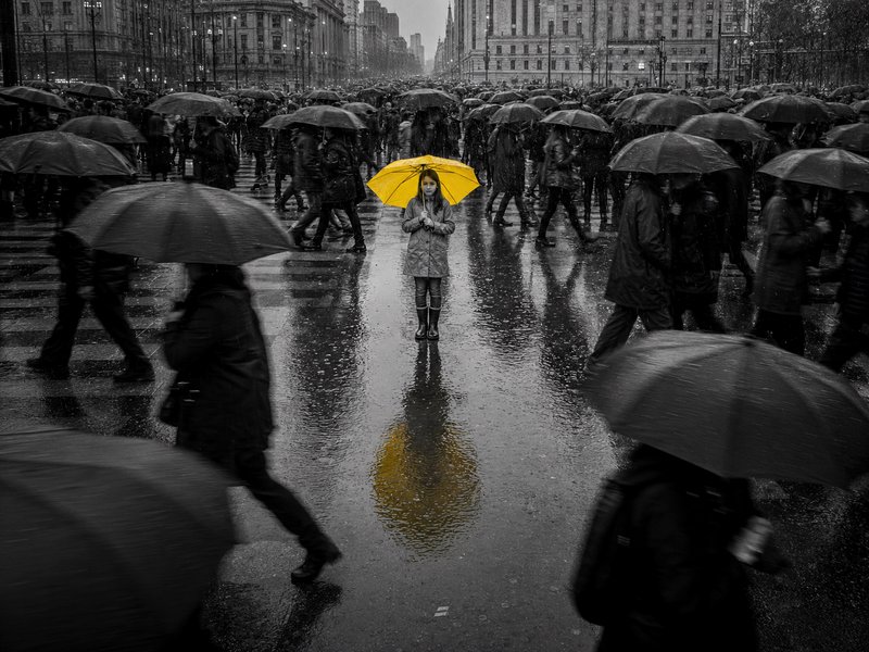 Lone woman with bright yellow umbrella stands still at the center of a monochrome rainy city crowd of pedestrians
