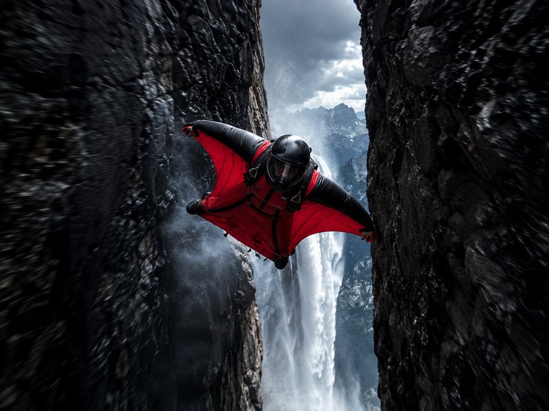Wingsuit flyer in red-black gear glides at high speed through a narrow rocky canyon beside a cascading mountain waterfall.