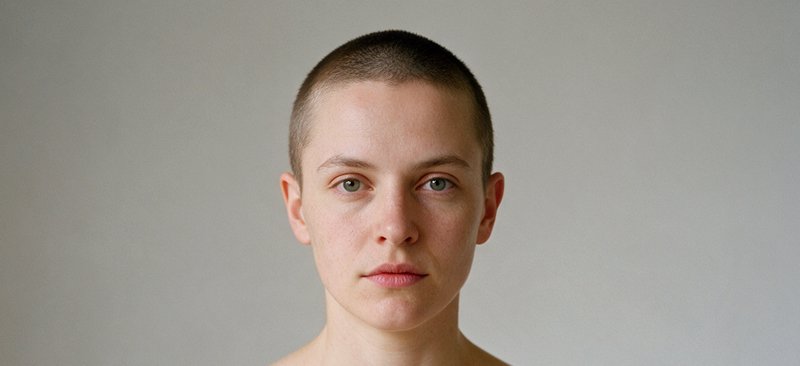 Close-up portrait of a person with a short buzz cut gazing directly at the camera against a plain...