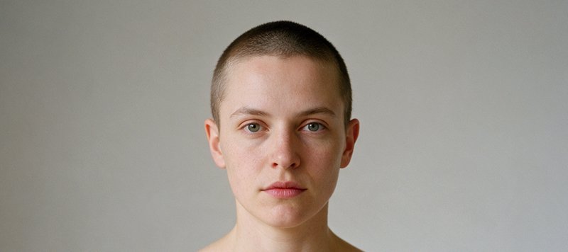 Close-up portrait of a person with a short buzz cut gazing directly at the camera against a plain...