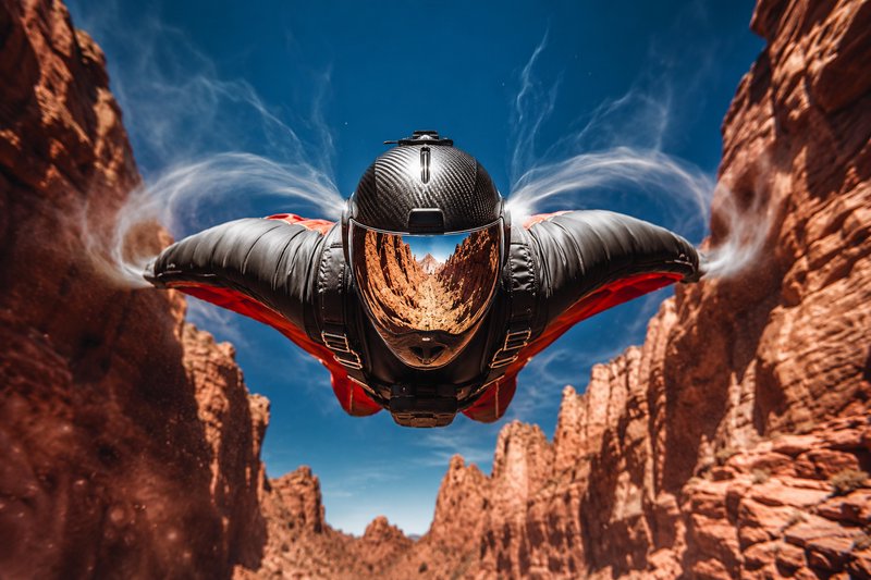 Wingsuit flyer glides head-on through a red rock canyon, wispy air vortex trails curl from their suit, reflective helmet v...