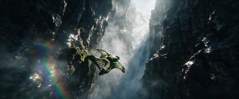 [Wingsuit flyer glides through a narrow misty rocky canyon beside a cascading waterfall with rainbow light flare]