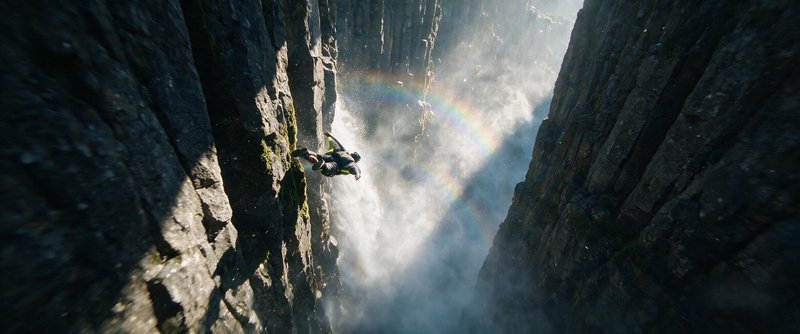 [Wingsuit flyer glides between steep canyon cliffs beside a misty waterfall, with a rainbow glowing through sunlit water s...