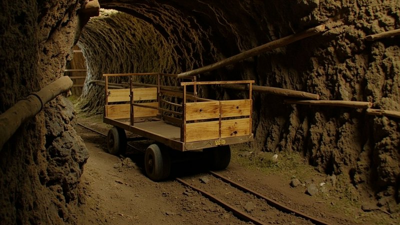 Weathered wooden mine cart rests on metal tracks inside a rough earthen underground mining tunnel...