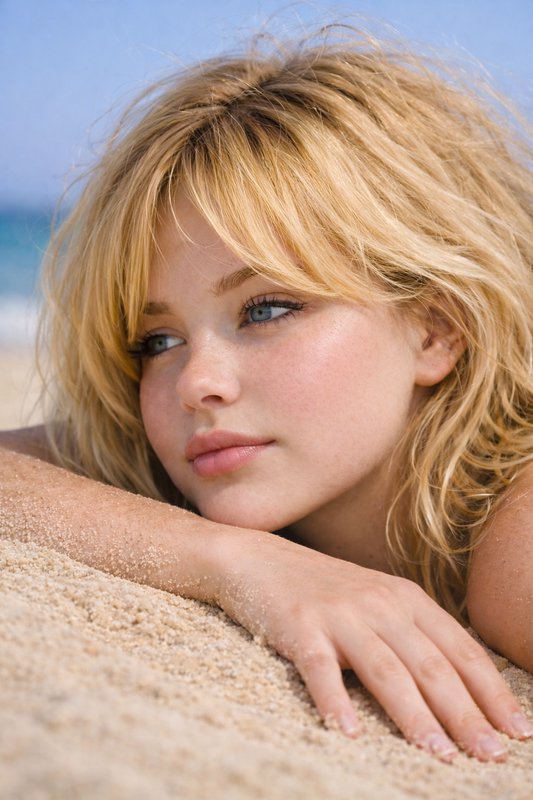 Young blonde woman with blue eyes rests on beach sand, gazing thoughtfully toward the ocean