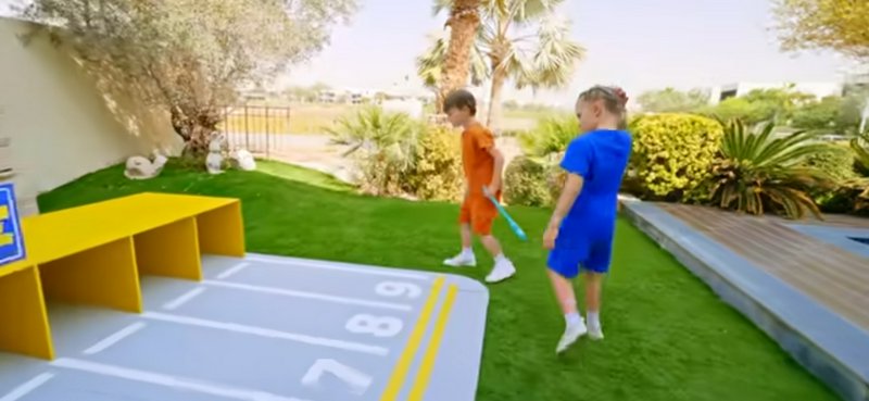 Two young children play near a numbered outdoor activity mat on a sunny residential artificial lawn