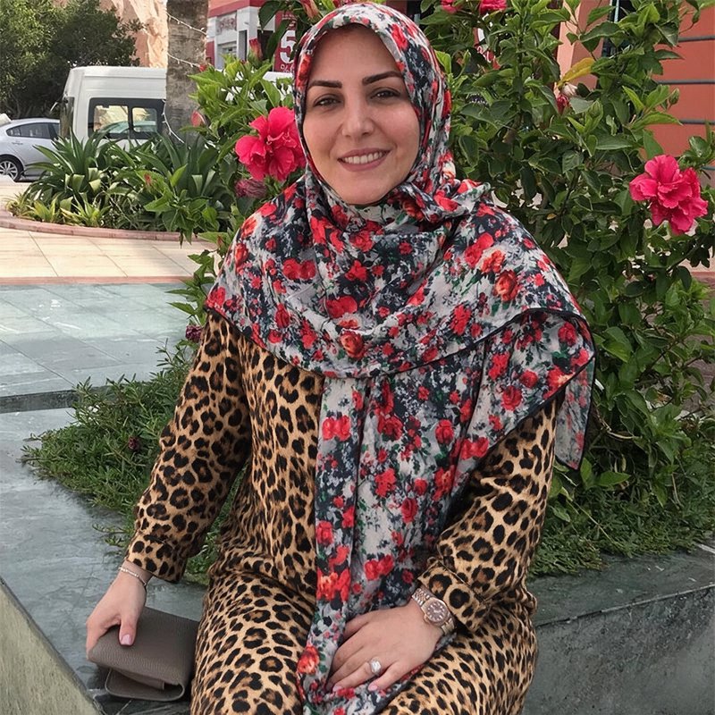 Smiling woman in a floral hijab and leopard print outfit sits outdoors beside pink hibiscus bushes