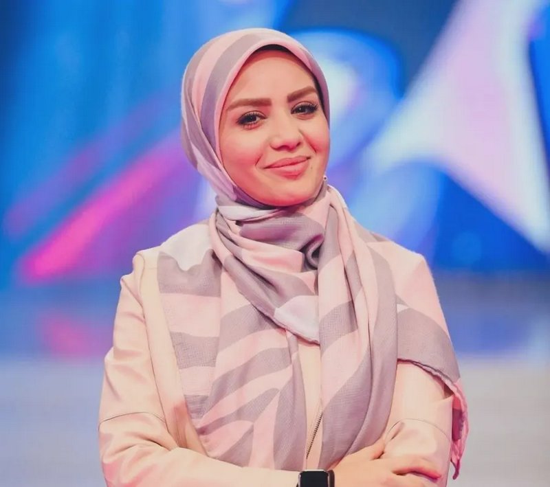 Smiling woman in a pastel striped hijab stands with crossed arms on a lit television studio set