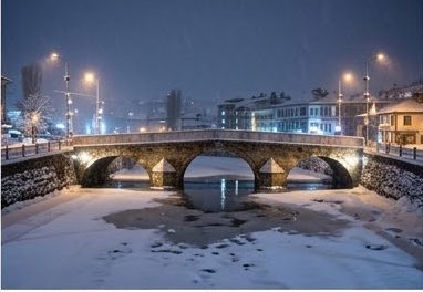 Historic stone arch bridge spans a partially frozen snowy river in a foggy winter town at night