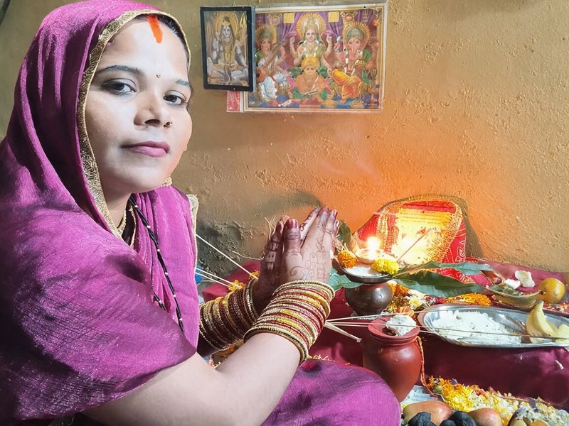 Indian woman in magenta headscarf holds prayer pose at a home Hindu puja altar with ritual offerings