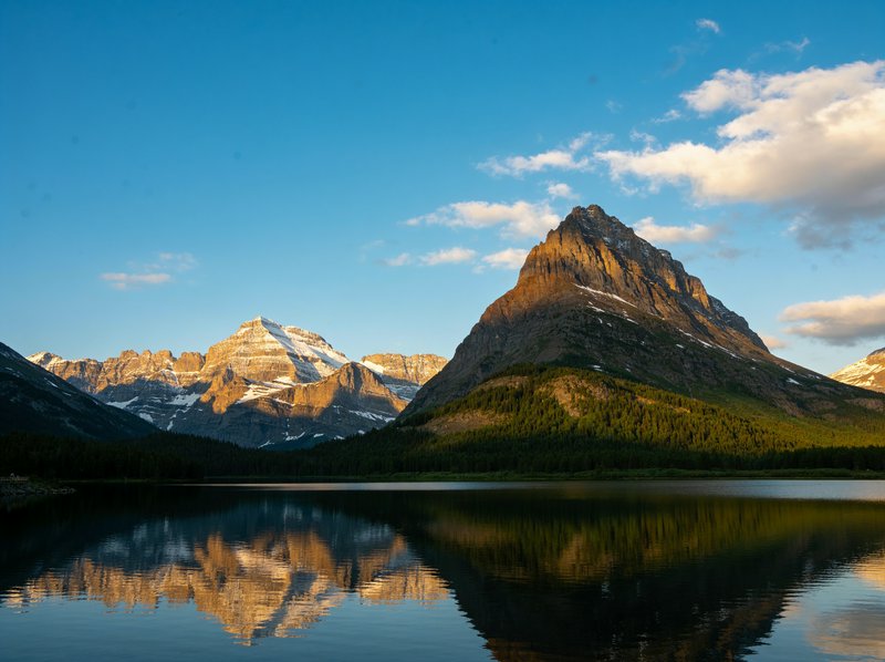 Sunlit jagged mountain peaks reflect in a still calm alpine lake under a bright blue sky
