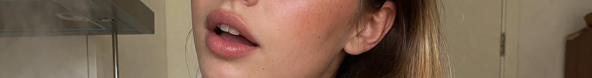 Extreme close up cropped view of a woman's dewy freckled cheek, glossy nude lips, and blonde hair