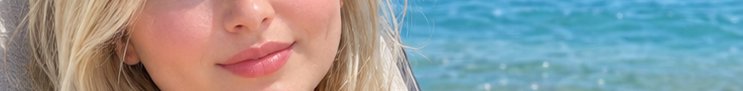 Cropped closeup of a blonde woman's lower face with a soft smile beside bright turquoise sunlit o...
