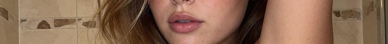 Extreme closeup of a woman's glossy plump lips, with wavy blonde hair against a beige tiled bathr...