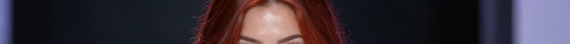 Tight cropped closeup of a woman's glossy red hair, dewy forehead, and arched brows against a dar...