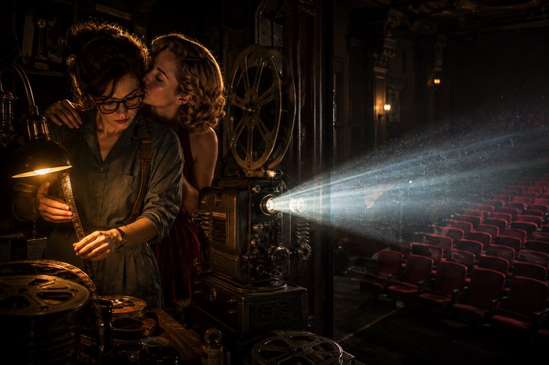 Two women in a vintage theater projection booth, one kisses the other as an antique film projecto...