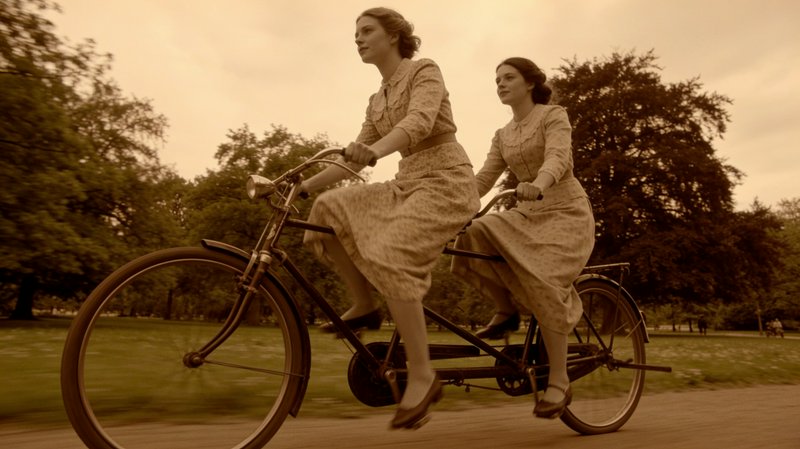 Sepia toned shot of two women in matching vintage dresses riding a tandem bicycle through a tree-lined park