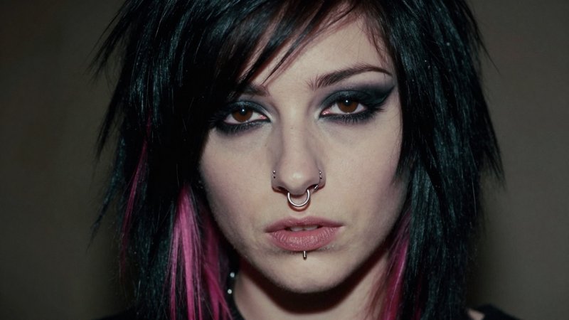 An ultra-realistic, ultra-detailed tight face portrait of a 20-year-old expressive woman from the late 90s and early 2000s emo subculture. She has dramatic, heavy black smudged eyeliner, intense dark eyeshadow, and thin, sharp penciled eyebrows. Her hair is a choppy, asymmetrical jet-black bob with long, sweeping side bangs covering one eye, featuring subtle neon pink streaks. Her expression is melancholic yet deeply intense, looking directly into the camera. She wears a small silver nose hoop and a vertical labret piercing. Sharp focus on the texture of her skin, individual eyelashes, and the fine strands of her hair. Shot on a 35mm film camera with a subtle grain, moody cinematic lighting with deep shadows, and an authentic, raw, slightly faded aesthetic of a vintage analog photograph.