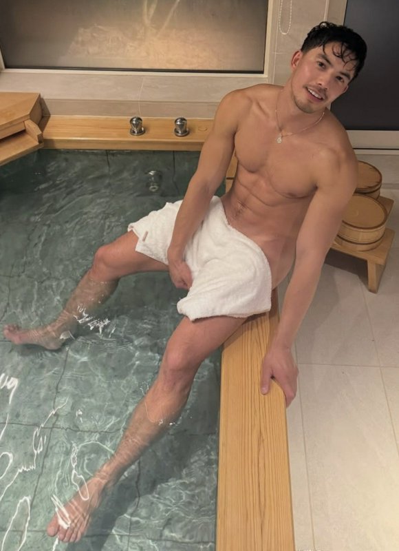 Shirtless man relaxing on the edge of an indoor spa pool with a white towel wrapped around his wa...