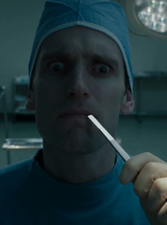 Close up of wide-eyed surgeon in blue scrubs holding a scalpel near his face in a dim operating room