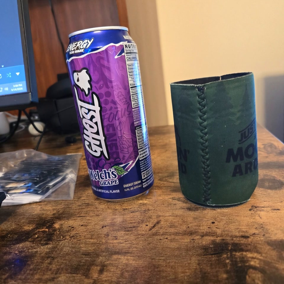 Purple Ghost Welch's Grape energy drink can sits next to a dark green stitched koozie on a rustic...