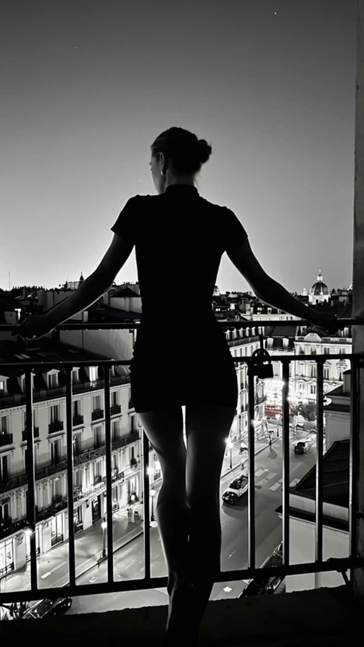 Silhouette of a woman standing on a balcony overlooking a lit city street at night, in black and ...