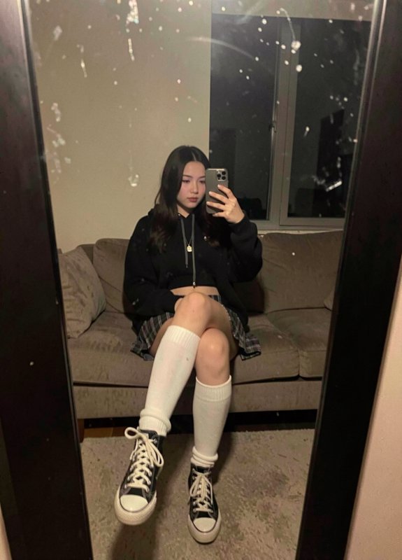 Teen girl taking a mirror selfie in a cozy living room wearing a black hoodie, plaid skirt, white...