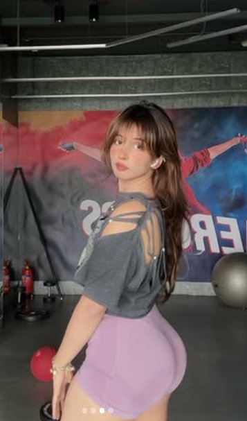 Young woman looks back over her shoulder posing in a gym, wearing a strappy grey top and lavender...