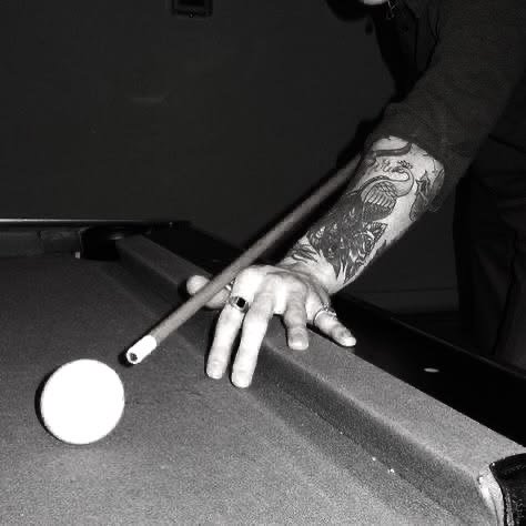 [Black and white close-up of a tattooed player holding a pool cue, aiming to strike the cue ball on a billiards table]