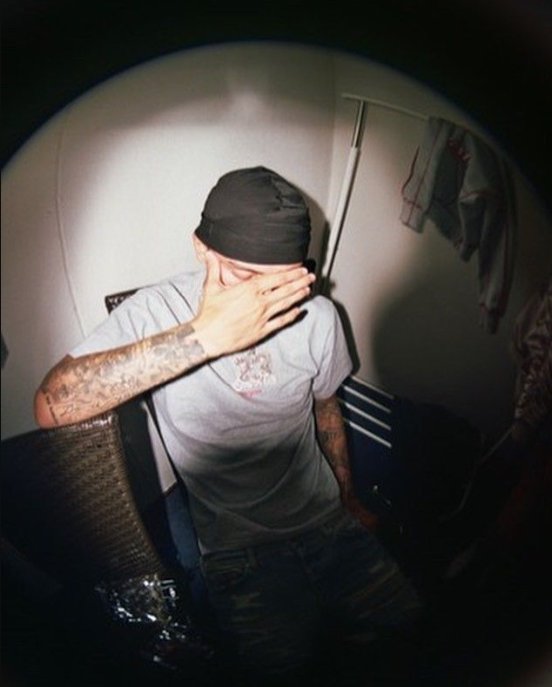 Grainy fisheye candid of tattooed man in a black durag covering his face with his hand, seated in...