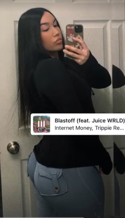 Young woman taking a casual mirror selfie with a Spotify now-playing widget overlay for the track...