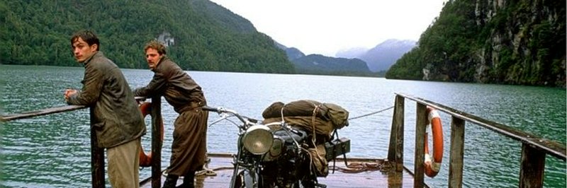 Two men stand on a wooden ferry with a packed vintage motorcycle crossing a forested mountain lak...